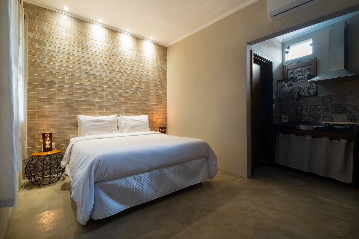 A bedroom with a large bed, a brick-patterned wall, a small bedside table with a lamp, and a kitchenette area with a window above the stove.