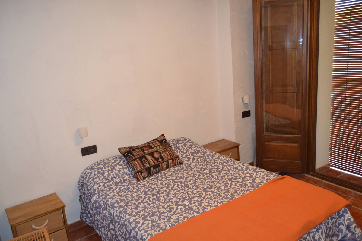 Une chambre à coucher comprenant un lit, une table de chevet, une armoire en bois et une fenêtre avec des stores.