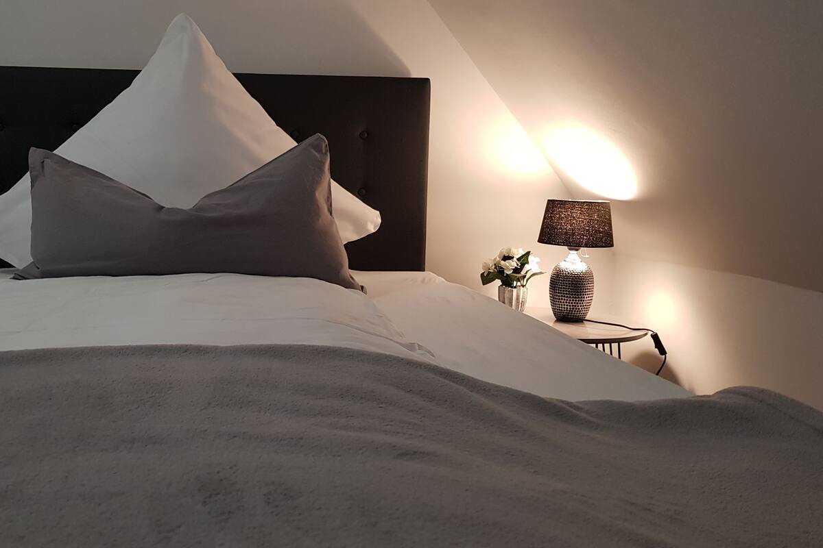 Un lit bien fait, avec une tête de lit sombre, une lampe de chevet et un petit vase avec des fleurs.