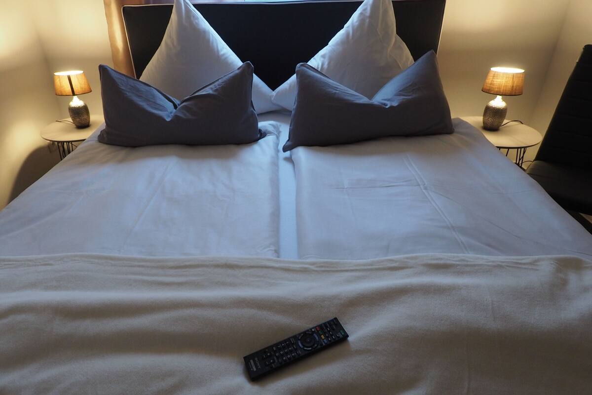 Un lit bien fait, avec des draps blancs, deux oreillers et une télécommande posée sur le lit. Deux lampes de chevet sont placées de chaque côté du lit.