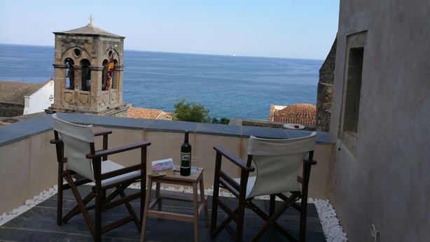 Ein Balkon mit zwei Holzstühlen und einem kleinen Tisch, der Blick auf Meer und einen historischen Turm bietet.