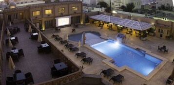 Un área de piscina en la azotea con sillones, mesas y una pantalla grande.