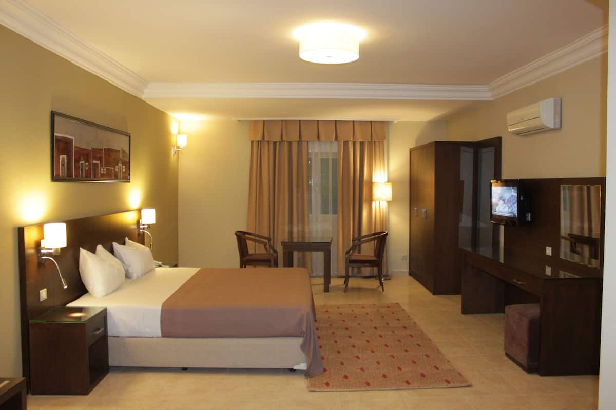 A hotel room with a bed, bedside tables, a desk with chairs, a television, and a large window with curtains.