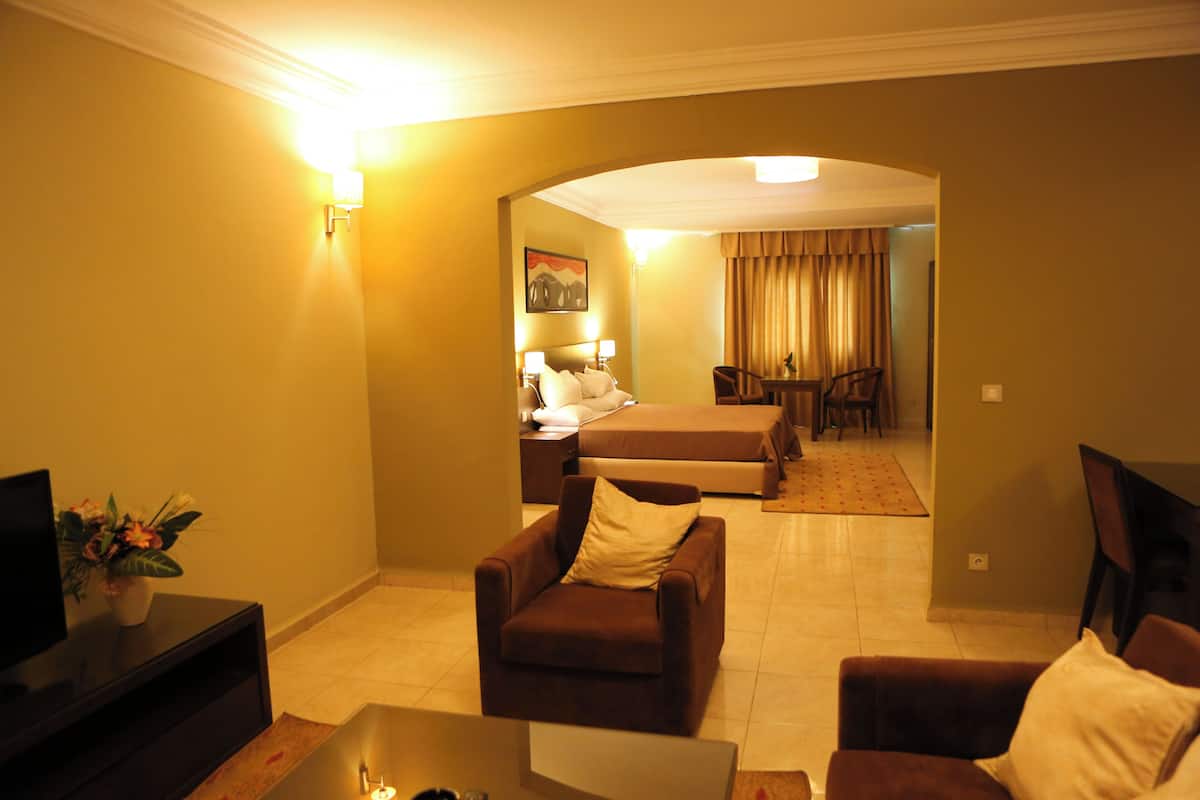 A hotel room with a sofa, armchair, and a small table. There is a bed in the background, a painting on the wall, and a lamp on the side table.