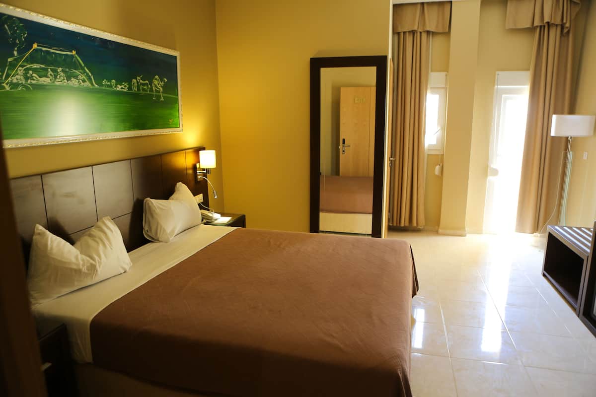 A hotel room with a large bed, a nightstand, a lamp, a painting on the wall, and a door leading to another room.