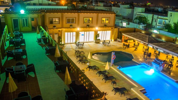 Un área de piscina en la azotea con sillones y sombrillas, rodeada por un edificio con ventanas amplias y suelo de baldosas.