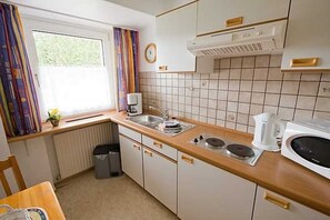 A compact kitchen with a sink, stove, and microwave. There is a window with curtains, a radiator, and a clock on the wall. - Apartment 1 - Panorama house (Sankt Andreasberg)