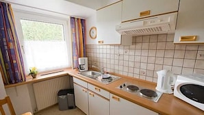 A compact kitchen with a sink, stove, and microwave. There is a window with curtains, a radiator, and a clock on the wall. - Apartment 1 - Panorama house (Sankt Andreasberg)