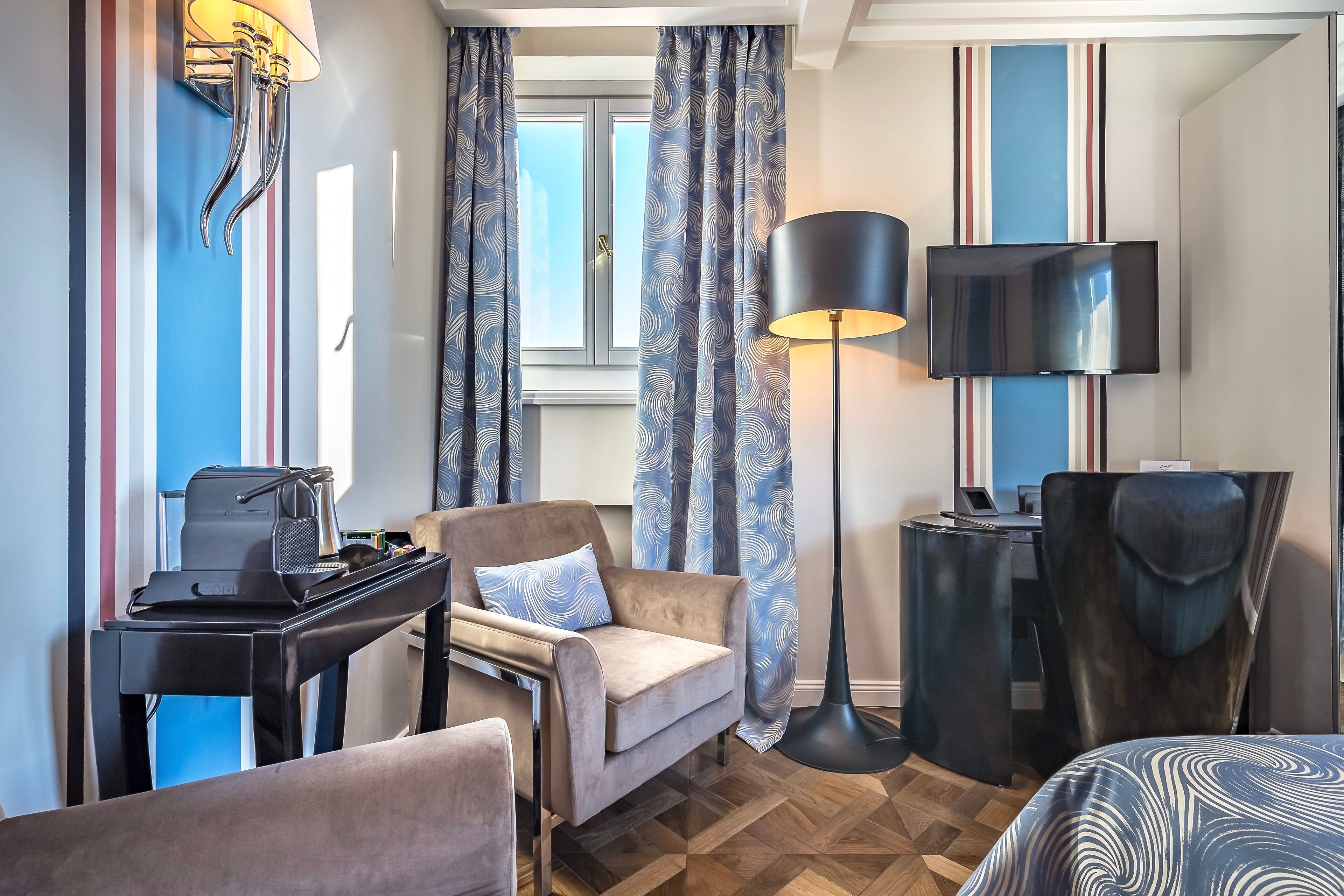 A hotel room with a bed, a desk with a computer, a chair, a lamp, and curtains.
