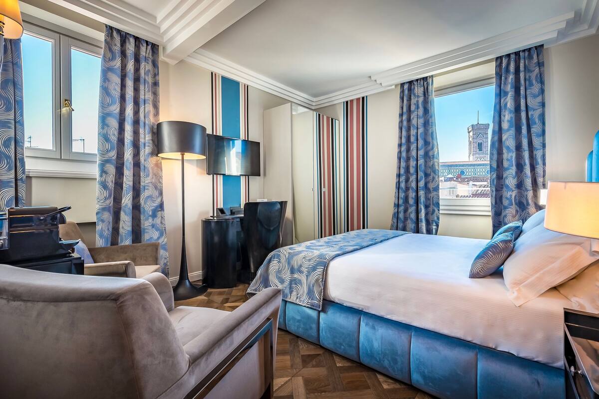 A hotel room with a bed, a desk, a chair, a lamp, and a view of a tower through the window.