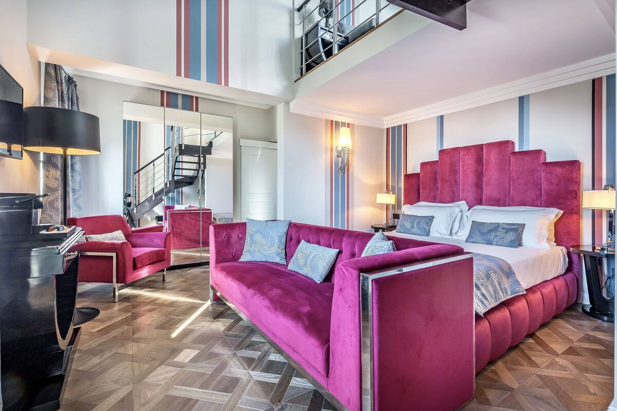 A modern hotel room with a large bed, a red sofa, a desk, and a staircase.