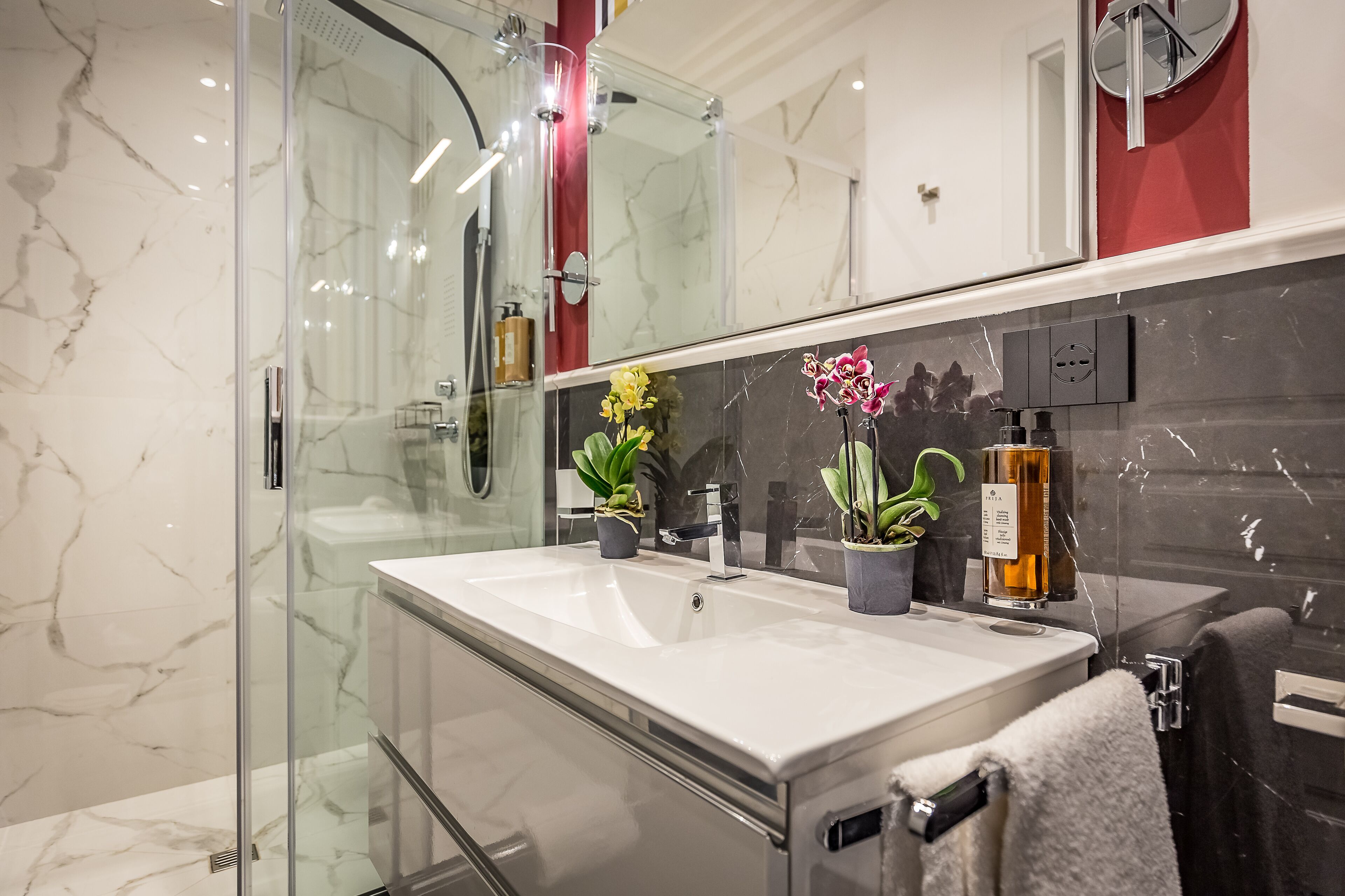 A modern bathroom with a glass shower enclosure, a large mirror, and a white sink with two potted plants.