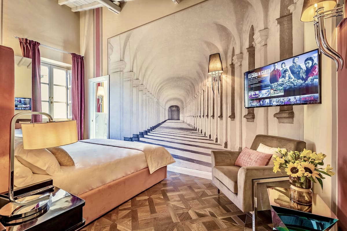 A hotel room with a large bed, a sofa, a TV mounted on the wall, and a hallway leading to a staircase.