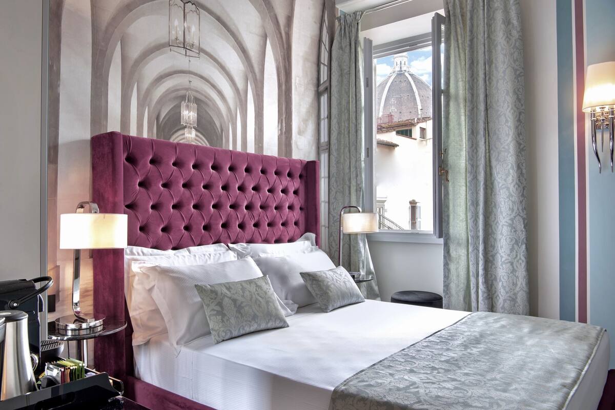 A hotel room with a large bed, a tufted headboard, a nightstand with a lamp, and a view of a building through the window.