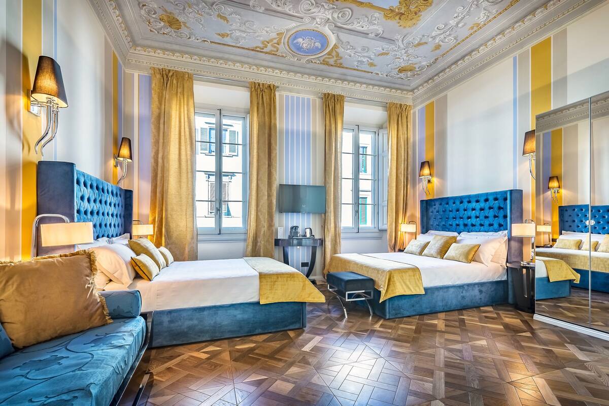 A luxurious hotel room with two beds, a sofa, a desk, and a large mirror.