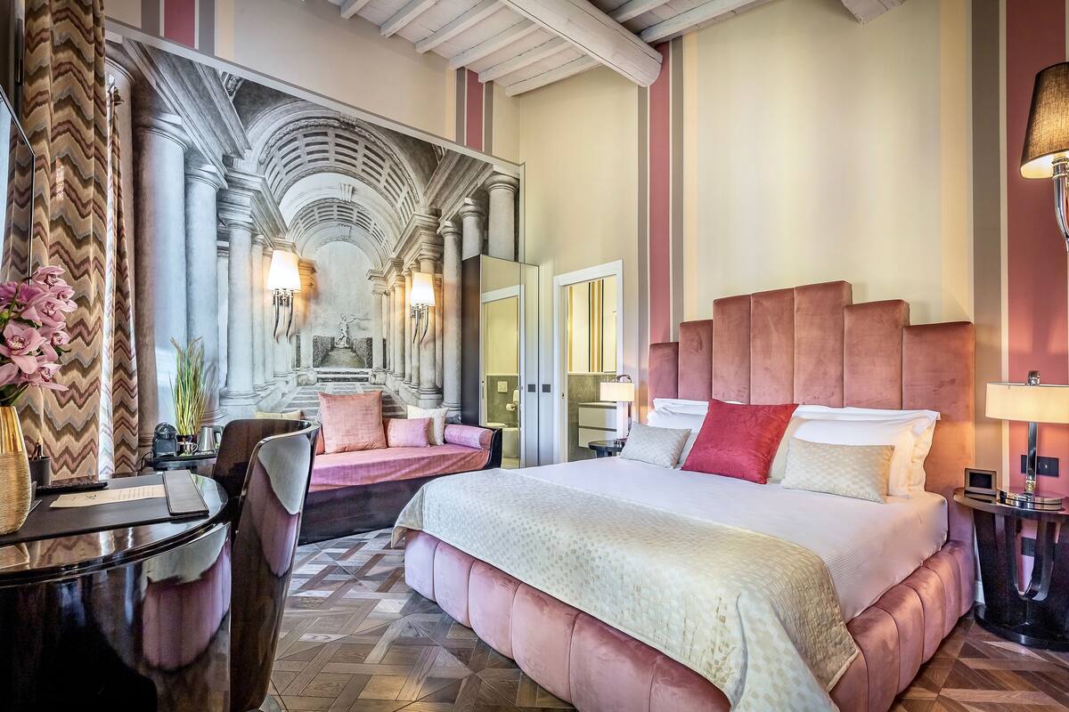 A hotel room with a large bed, a sitting area with a sofa, a dining table, and a decorative mirror.