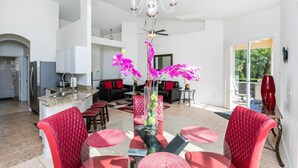 A spacious living area with a dining table, red upholstered chairs, a chandelier, and a kitchen in the background.