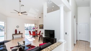 A spacious living room with a large TV, comfortable seating, a dining area with red chairs, and a view of the outdoors.