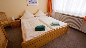 Ein Einzelbett mit Holzrahmen, zwei Kissen, ein grünes Handtuch, ein Nachttisch mit Lampe sowie ein gerahmtes Bild an der Wand.