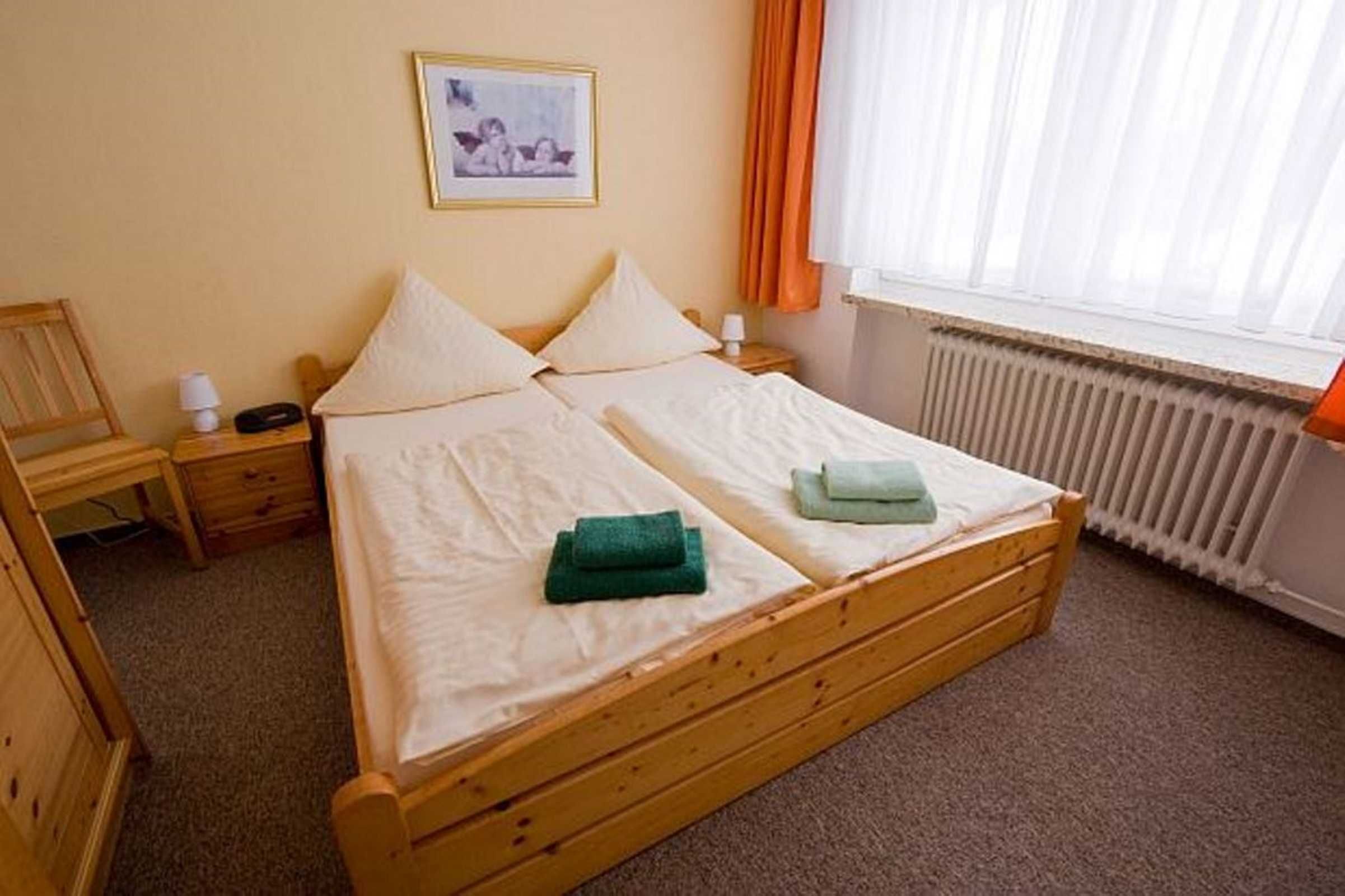 A single bed with a wooden frame, two pillows, a green towel, a bedside table with a lamp, and a framed picture on the wall.