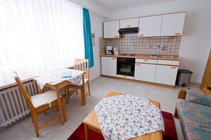 Une cuisine compacte avec des meubles blancs, une machine à café et un petit four. On y trouve une table à manger avec des chaises, ainsi qu’un canapé dans l’espace salon.