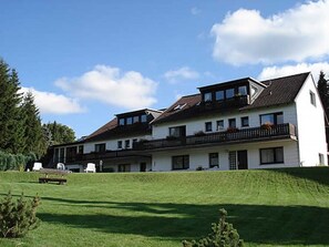 A two-story building with a sloped roof, multiple balconies, and a grassy front lawn. - Apartment 4 - Panorama of the house (Sankt Andreasberg)