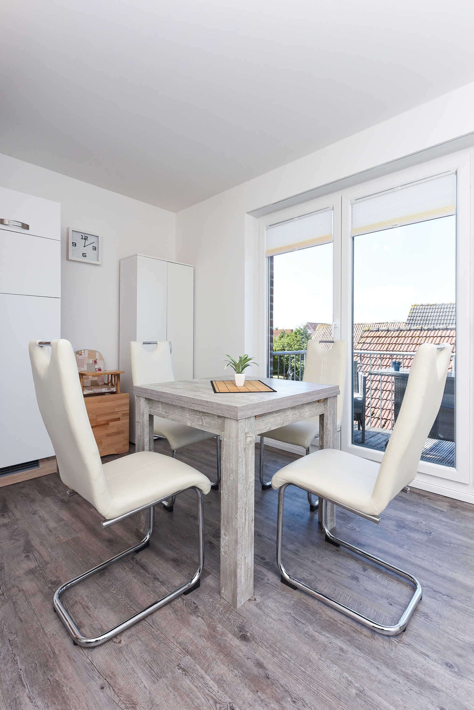Une cuisine moderne avec une table en bois et deux chaises blanches, un meuble blanc et une fenêtre donnant sur des immeubles.