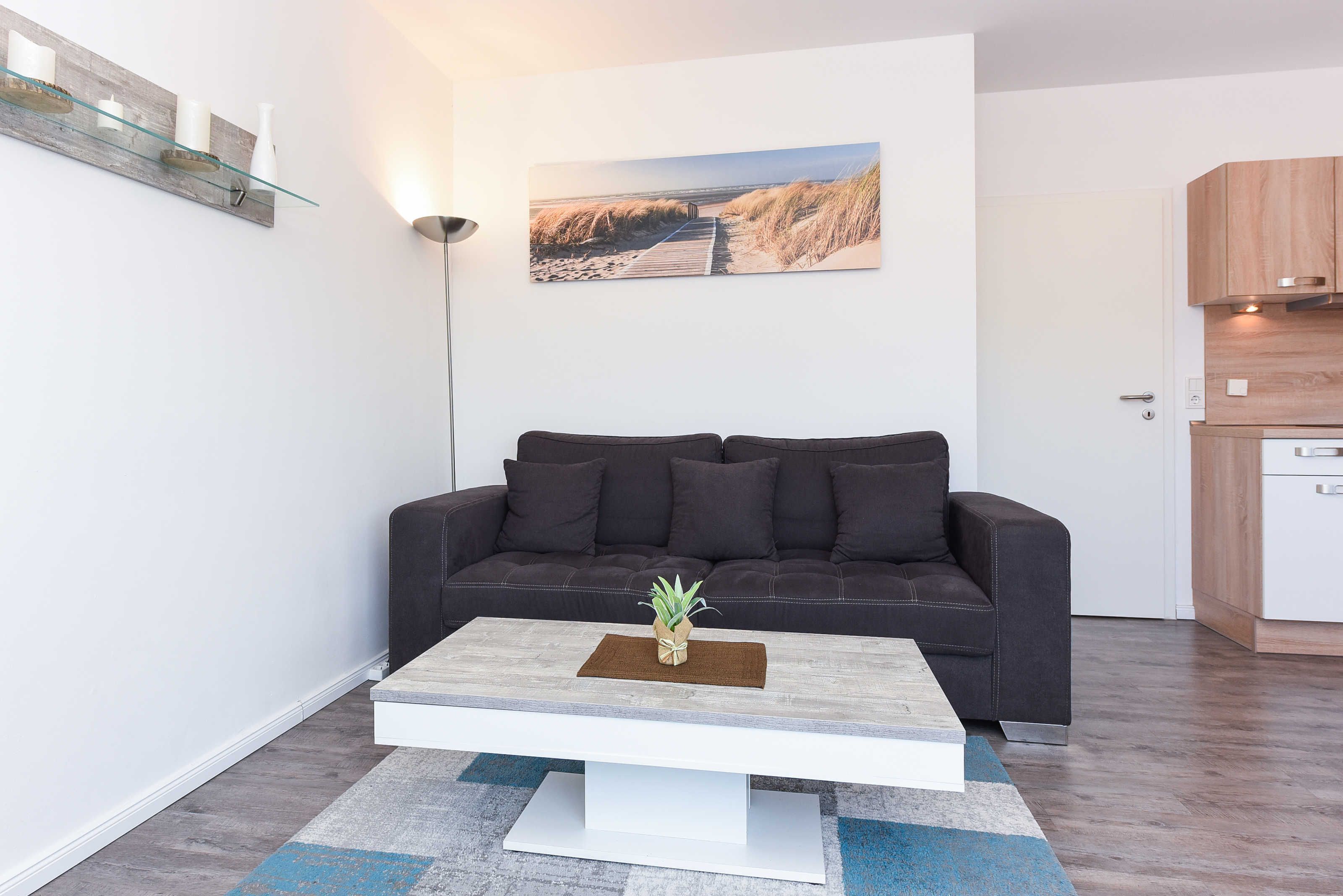 Una sala de estar moderna con un sofá oscuro, una mesa de centro blanca con una planta y un cuadro con temática playera en la pared.
