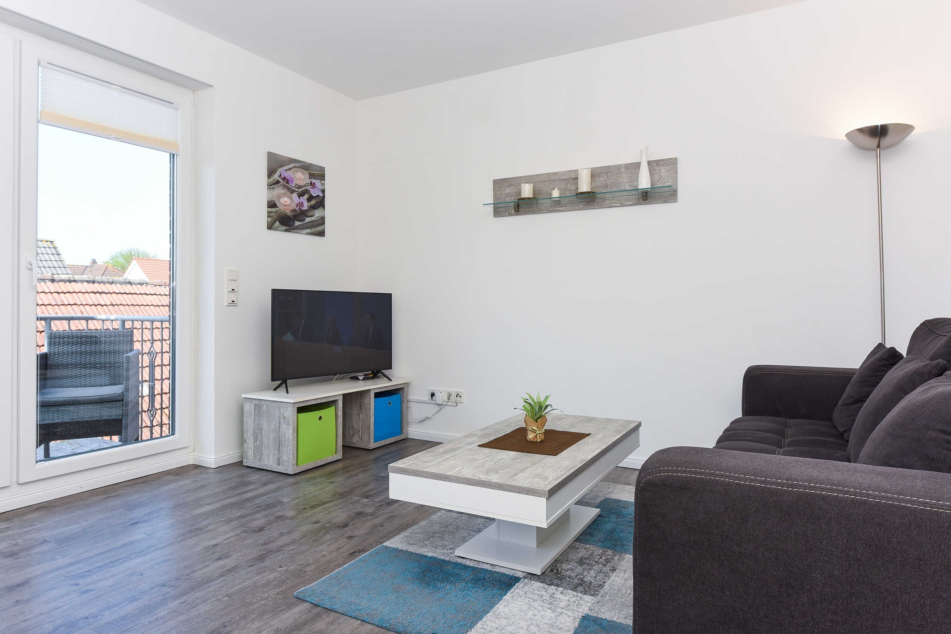 A modern living room with a flat-screen TV, a coffee table with a plant, a grey sofa, and a large window with a view of buildings.