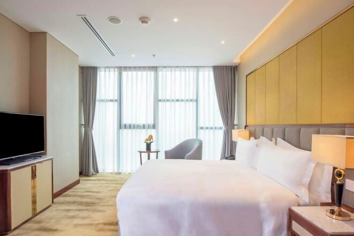 A hotel room with a large bed, a flat-screen TV, a small table with a lamp, and a view of the outside through large windows.