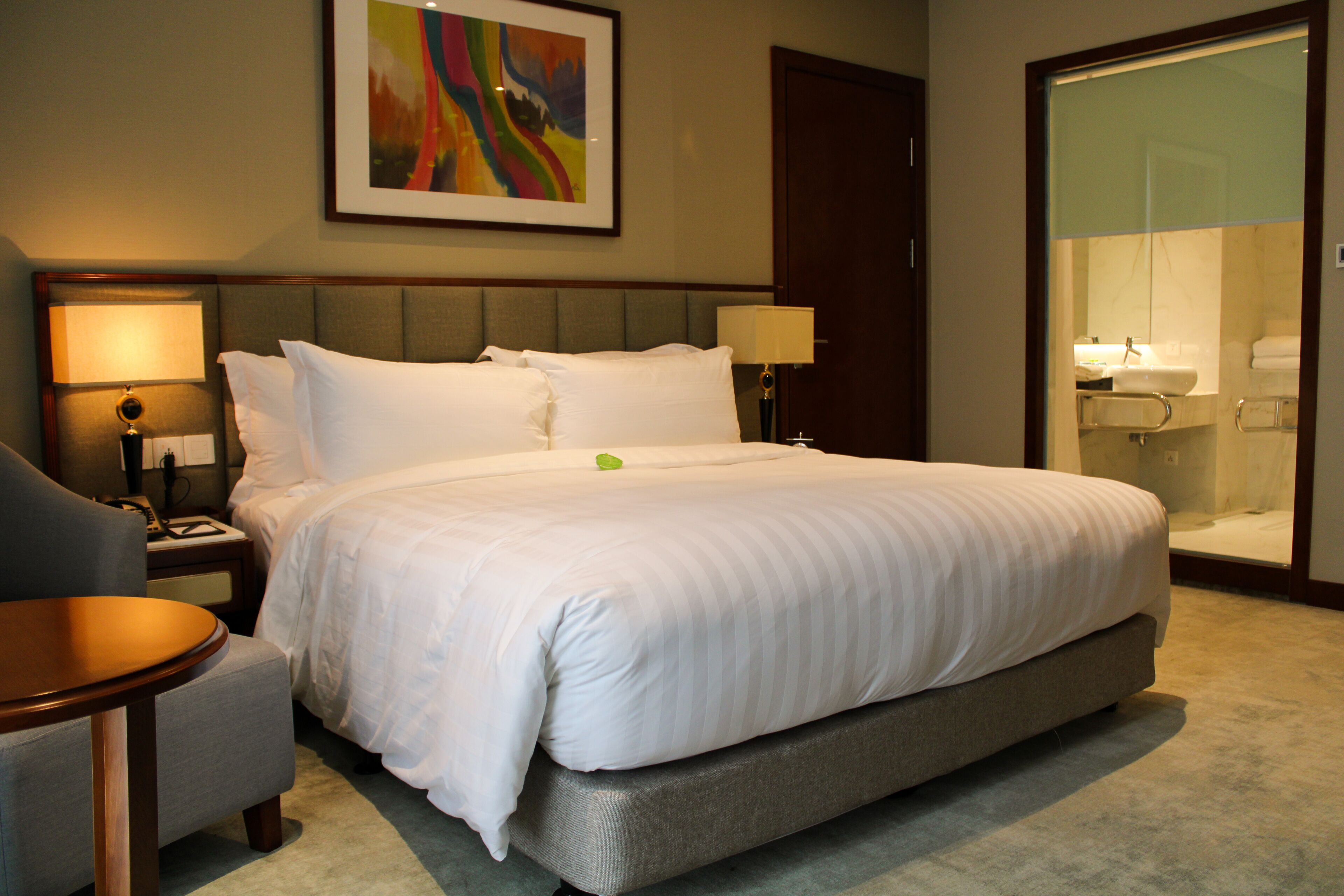 A hotel room with a large bed, a bedside table, a chair, a desk, and a bathroom visible through a glass door.