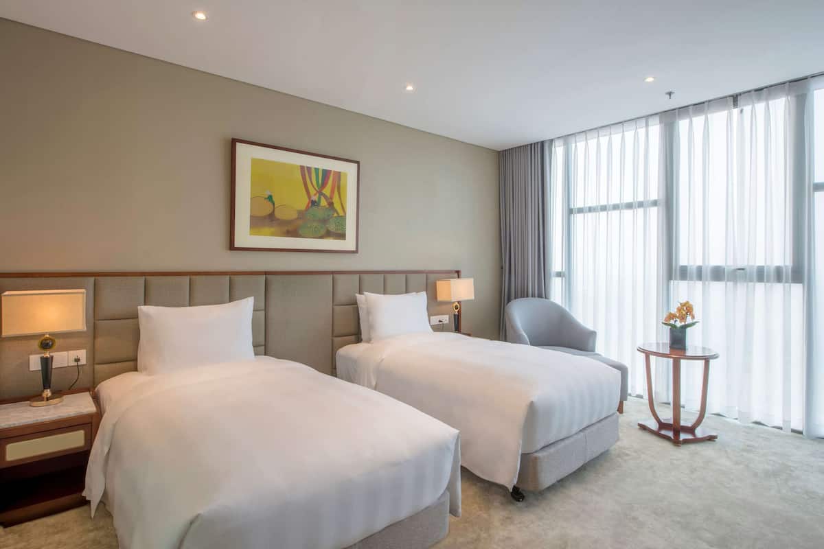 Habitació d'hotel amb dos llits, un capçal, una tauleta de nit amb una làmpada, una cadira, una taula petita amb un vas i una finestra amb cortines.