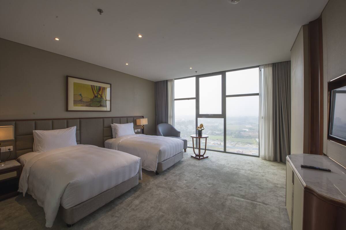 Habitació d'hotel amb dos llits, una finestra gran amb cortines, un quadre a la paret i una taula de nit amb una làmpada.