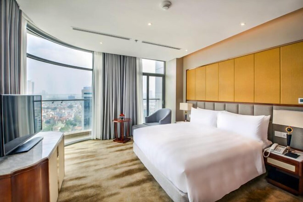 A hotel room with a large bed, a desk with a flat-screen TV, a chair, a nightstand with a phone, and a view of the city through large windows.
