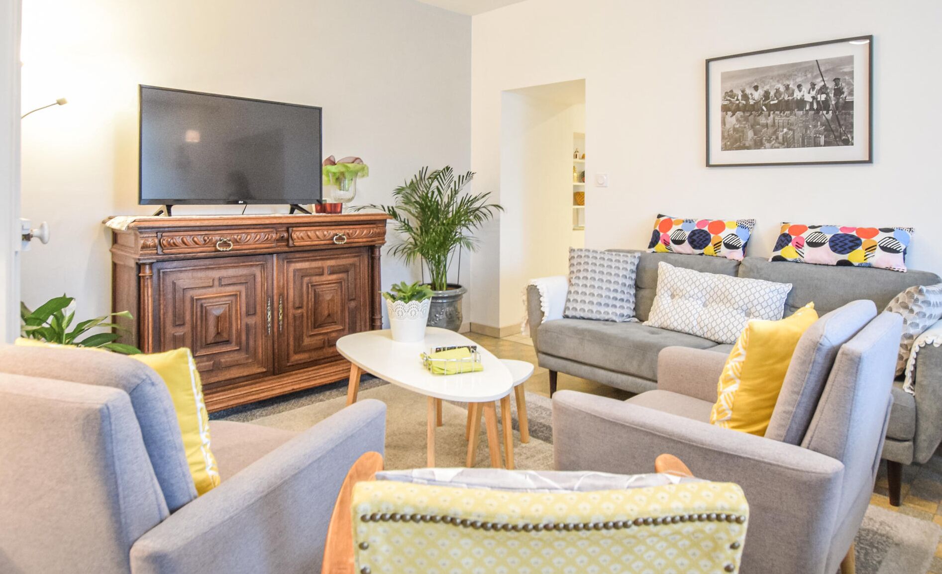 Ein Wohnzimmer mit einem Fernseher auf einer Holzkommode, einem grauen Sofa mit gemusterten Kissen und einem kleinen Tisch mit einer Pflanze.