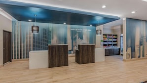 A reception area with a modern design, featuring a cityscape mural, wooden reception desks, and a well-stocked beverage fridge. - La Quinta Inn and Suites by Wyndham Houston Spring South (Spring)