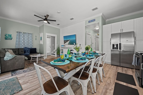 A modern kitchen with a stainless steel refrigerator, a dining area with a wooden table and white chairs, and a living room with a sofa and patterned curtains.