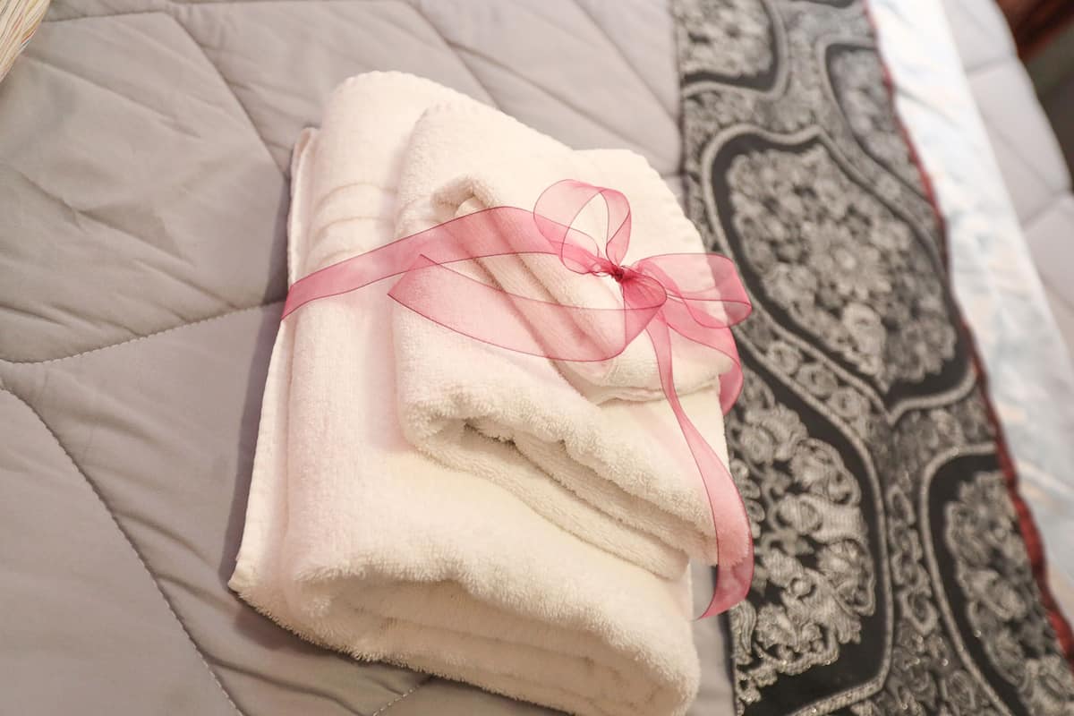 A neatly folded white towel with a pink ribbon tied around it, placed on a bed with a patterned bedspread.