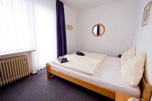 Un lit simple avec un cadre en bois, un miroir rond accroché au mur et un radiateur placé sous la fenêtre.