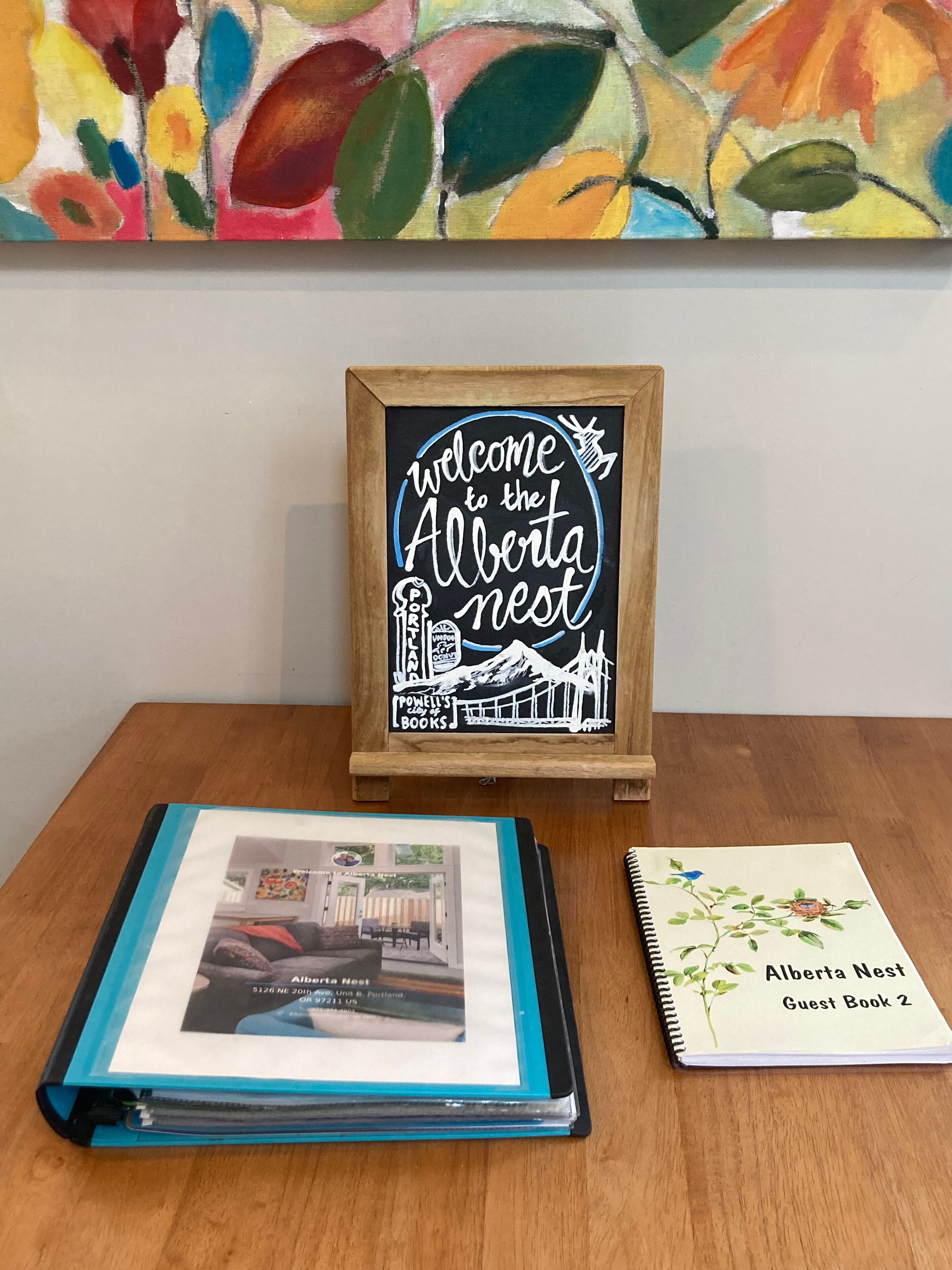A wooden sign with "Welcome to the Alberta Nest" and a photo of a building. Below, a blue folder and a spiral notebook with "Alberta Nest Guest Book 2" are on a wooden table.
