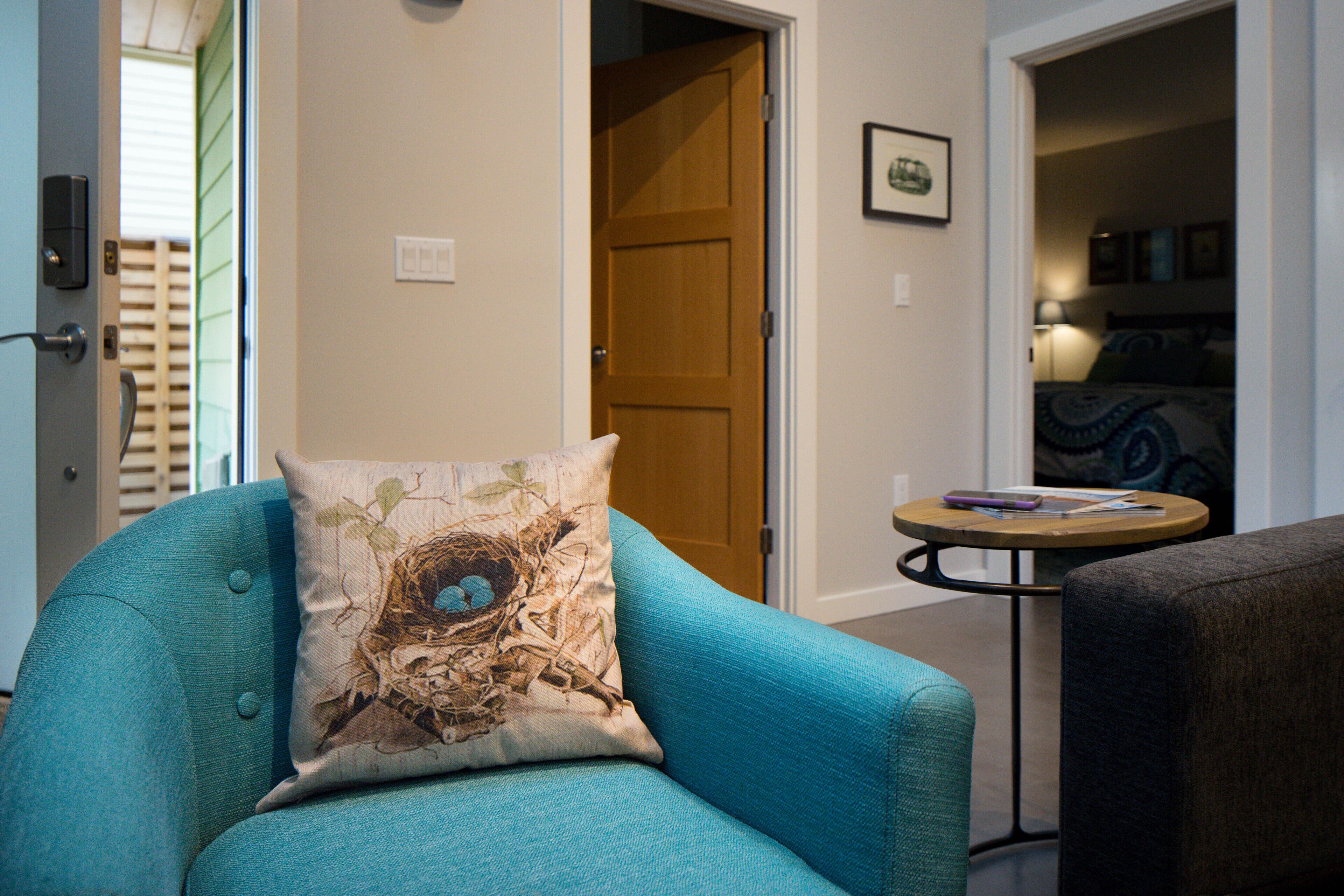 A teal armchair with a decorative pillow, a wooden door, and a small round table with a lamp in a hallway.