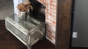 Ein Kamin aus Backsteinen mit einem dekorativen Metallornament darüber, ein Metallheizkörper darunter und eine Glasplatte mit einer Vase mit Trockenblumen.