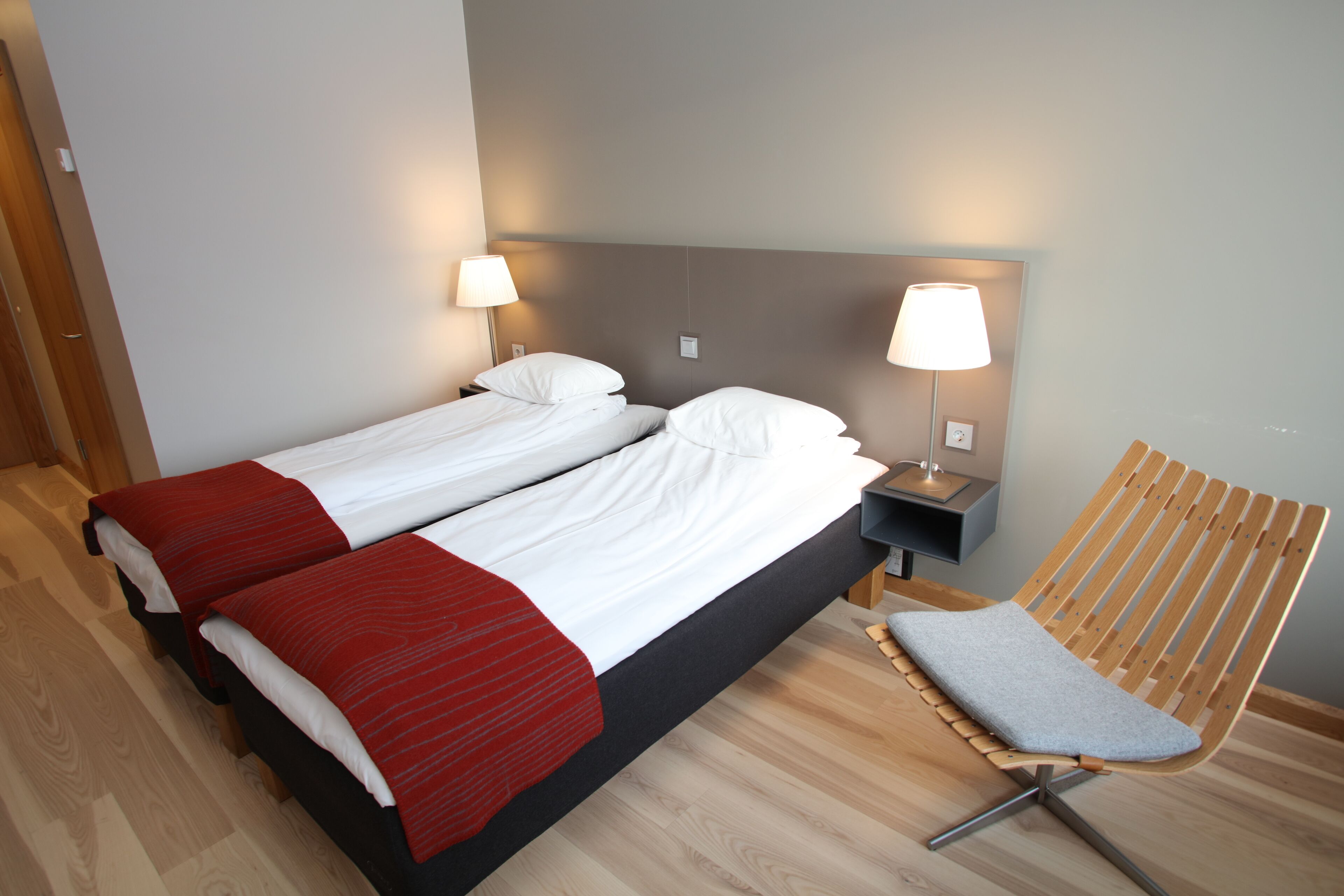 A double bed with white and red bedding, two bedside tables with lamps, a wooden chair, and a wooden floor.