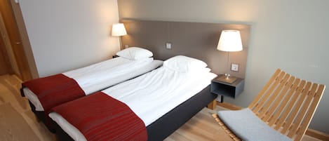Una cama doble con ropa de cama blanca y roja, dos mesitas de noche con lámparas, una silla de madera y suelo de madera.