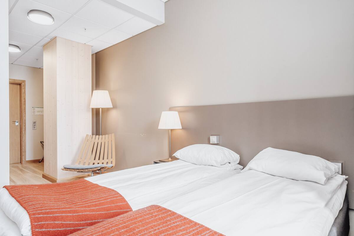 Una habitación de hotel con cama, dos lámparas de noche, una silla de madera y una manta roja.