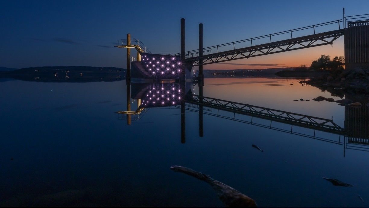 Un pont surmonté d’une grande structure illuminée, dont le reflet se mire dans l’eau.