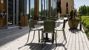 En træterrasse med udendørs møbler og en stor bygning med mange vinduer i baggrunden.