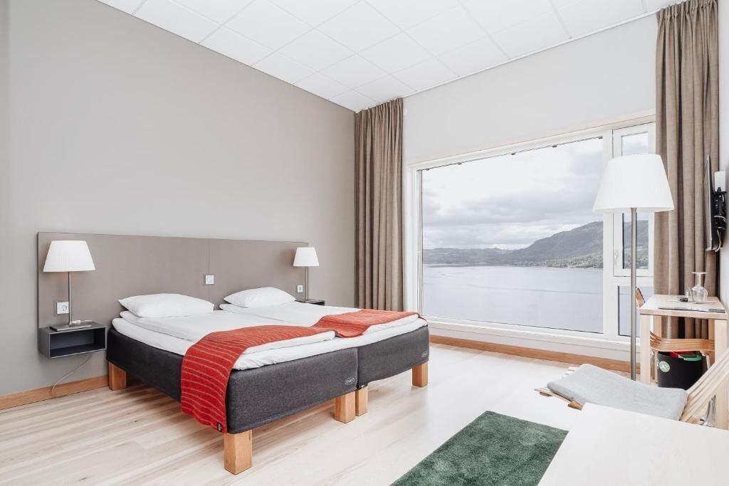 Una cama doble con una manta roja, dos mesitas de noche con lámparas, vista a un lago y un escritorio pequeño con una lámpara.