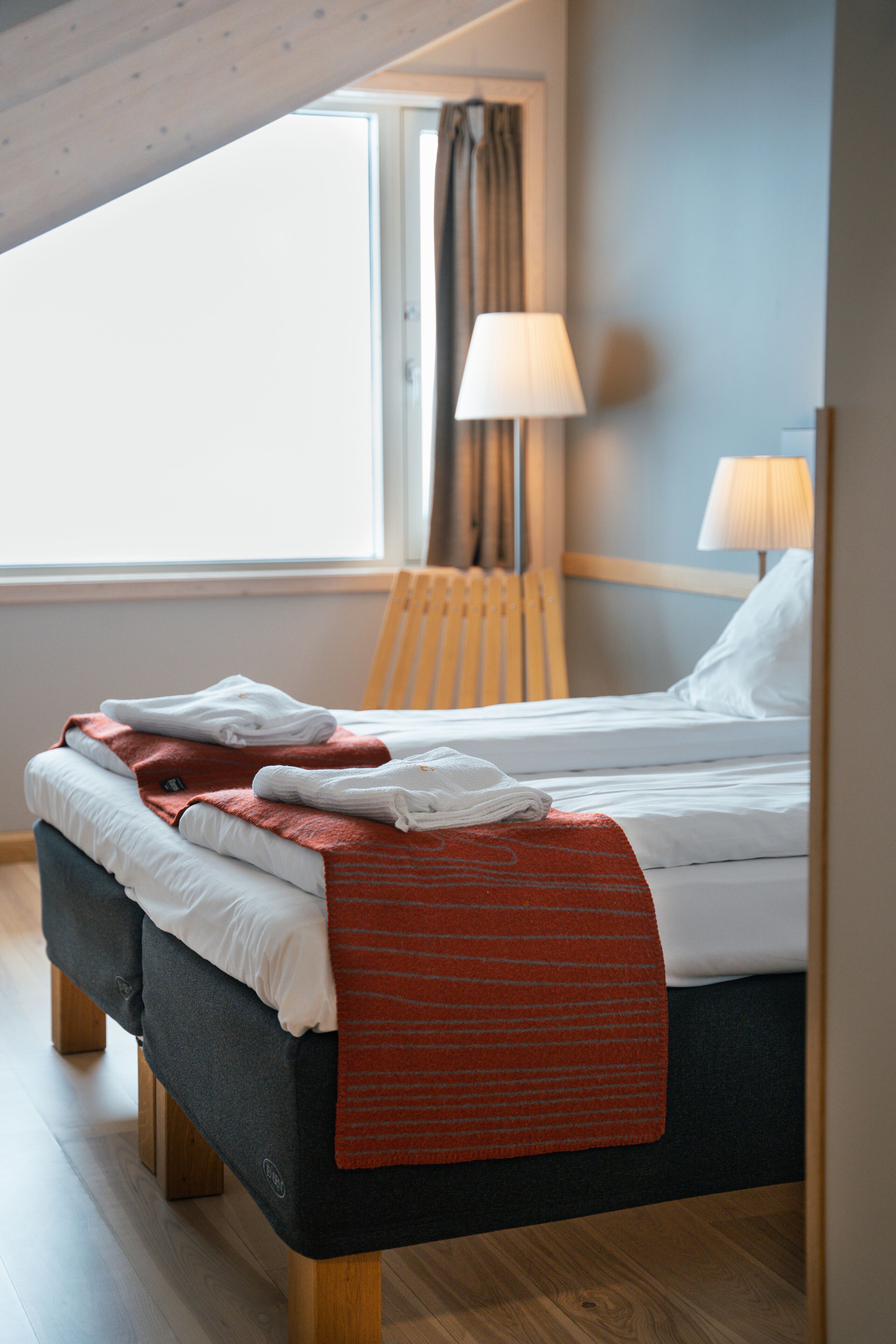 Una cama bien hecha con mantas blancas y rojas, una silla de madera y una lámpara a su lado.