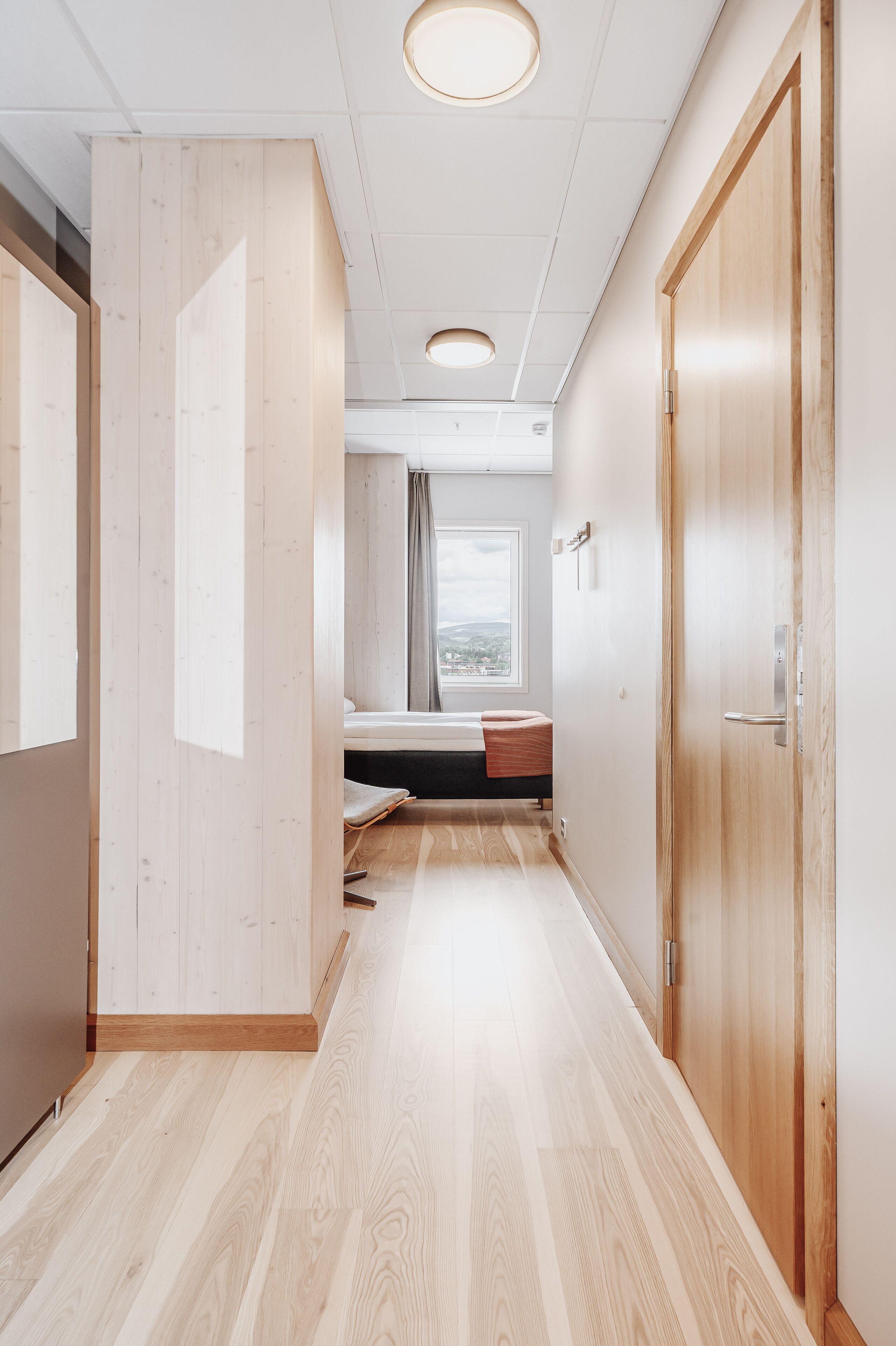 A modern hotel corridor with wooden flooring, a bed with a view, and a wooden door.
